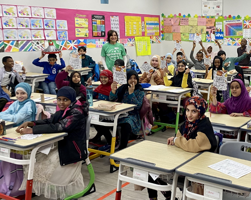 Classroom of early years students holding up agriculture books, with a volunteer in a green shirt standing and smiling among them